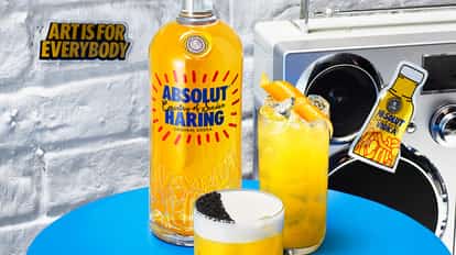 Absolut Haring Fizz and an Absolut Haring Collins sitting on a round blue table next to a bottle of Absolut Haring and a sticker on the wall saying 'Art is for everybody'-