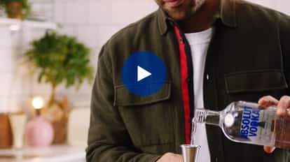 Rico pouring Absolut vodka from a bottle into a jigger over a mixing glass in a kitchen.
