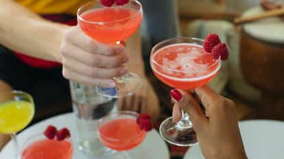 Several pink cocktails in coupe glasses garnished with raspberries, two hands clinking glasses.