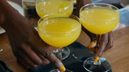 Three yellow sour cocktails in coupe glasses held by hands with an Absolut vodka bottle in the background.