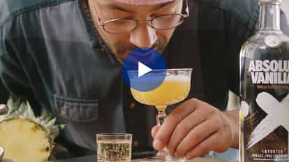 Rico smelling a yellow cocktail in a coupe glass with an Absolut Vanilla bottle on a wooden board