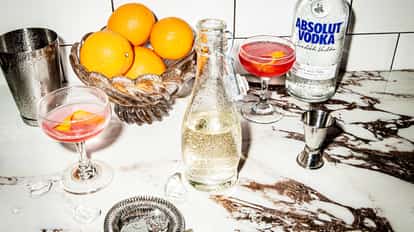 Triple Sec next to a bottle of Absolut Vodka and a Cosmopolitan cocktail, surrounded by fresh whole oranges.