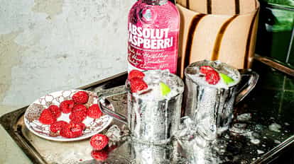 Two aluminum mule mugs of the Raspberry Vodka Mule made with Absolut Raspberri, a twist on a traditional Moscow Mule, filled to the top with ice, garnished with fresh raspberries, sitting beside a saucer of fresh raspberries.