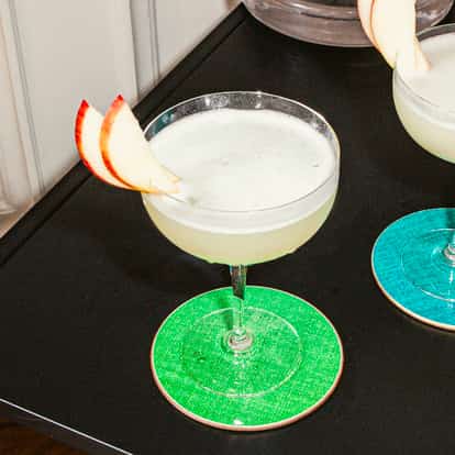 Two apple martinis garnished with fresh apple slices, served in coupe glasses on colorful coasters, placed on a black table.