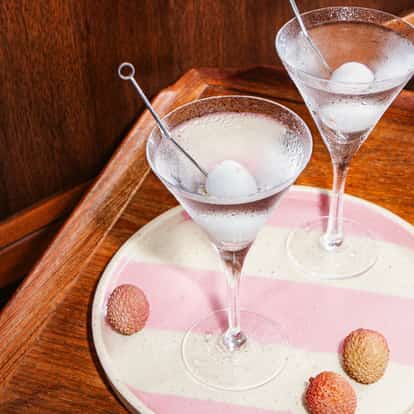 Zwei gekühlte Lychee-Martinis in Martinigläsern mit aufgespießten Litschis, serviert auf einem gestreiften Tablett mit frischen Litschis.