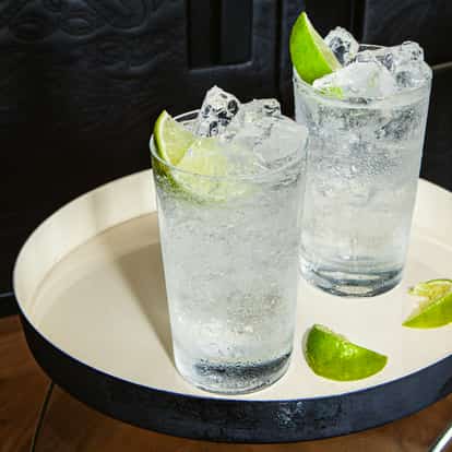 Zwei Gläser mit einem klaren, sprudelnden Cocktail mit Eis und Limettenstücken auf einem runden weißen Tablett, daneben weitere Limettenstücke. Die Umgebung ist modern mit Büchern und dunklem Hintergrund.