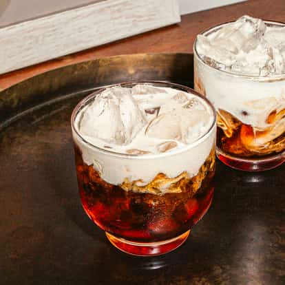 Zwei niedrige Gläser mit einem geschichteten Cocktail aus dunklem Likör, Eis und einer cremigen weißen Schicht, serviert auf einem dunklen Tablett auf einer Holzoberfläche. Das Getränk wirkt kühl und reichhaltig.