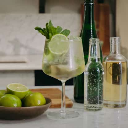 Hugo Spritz cocktail in a wine glass next to a bowl of fresh limes, elderflower liqueur and soda water.