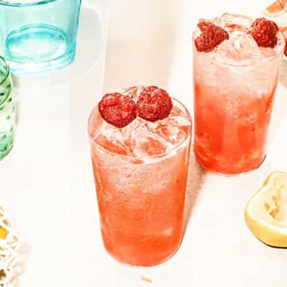 Two Raspberry Collins cocktails garnished with fresh raspberries surrounded by colored glasses.