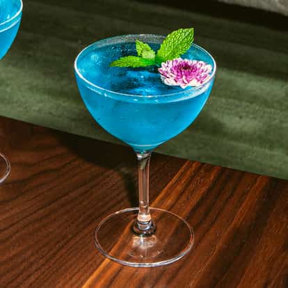 Koktajl Lotus Flower Martini podany w kieliszku typu coupe, w jaskrawoniebieskim kolorze, udekorowany różowym kwiatem i świeżą miętą, stojący na drewnianym stole.