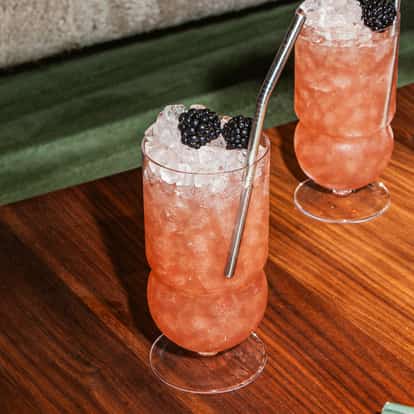 A-Veranda-Moment-Cocktail im hohen Glas mit Crushed Ice, Brombeeren und Metallstrohhalm, stehend auf einem Holztisch.