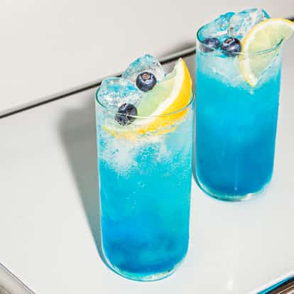 Blueberry Fizz Cocktails, serviert in hohen, gekühlten Gläsern, leuchtend blau und voller Eis, garniert mit Zitronenspalten und frischen Blaubeeren.