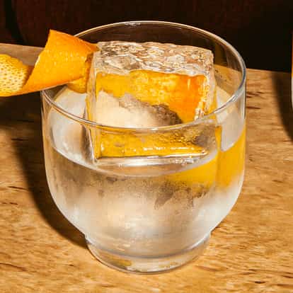 Coquetel Absolut Gangster gelado em copo redondo com grande cubo de gelo claro e casca de laranja sobre mesa de madeira, com castanhas de caju por perto.