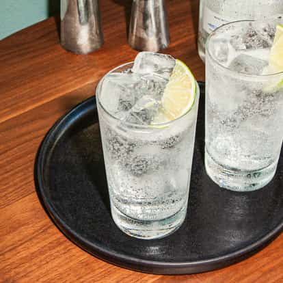 Two vodka soda cocktails with ice cubes and lime wedges served in highball glasses on a black tray on a wooden table.