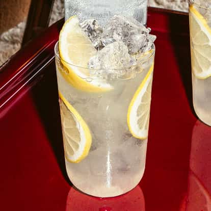 Dois copos altos de vodka com limonada, gelo e fatias de limão sobre mesa vermelha brilhante, drink cítrico refrescante.