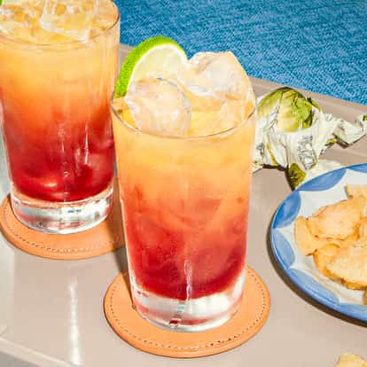Dwa koktajle Bay of Passion z czerwono-pomarańczowymi warstwami i limonką na tacy obok chipsów, na tle niebieskiej sofy.