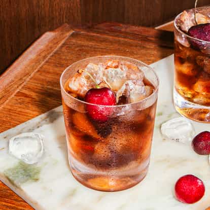 Due cocktail Black Russian con ghiaccio e ciliegie in bicchieri bassi su un vassoio di marmo, circondati da ghiaccio e ciliegie fresche.