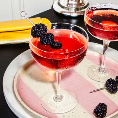 Two Blackberry Martini cocktails in coupe glasses with fresh blackberry garnishes on a pink striped tray beside a yellow clutch.