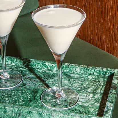 Dwa koktajle Blanc Martini w wysokich kieliszkach na zielonej marmurowej tacy, na tle zielonej sofy i drewnianej ściany.