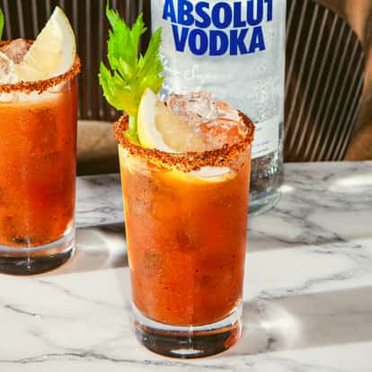 Dos cócteles Bloody Mary con limón, apio y borde especiado sobre mesa de mármol junto a plato de aceitunas verdes y botella de vodka.