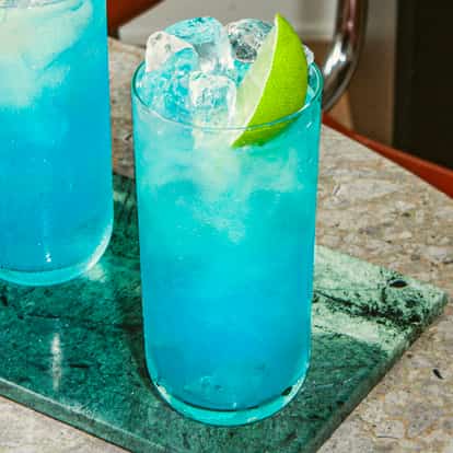Zwei hohe Gläser des Blue Andy Cocktails, leuchtend blau mit Eis und Limette auf einer Steinplatte.