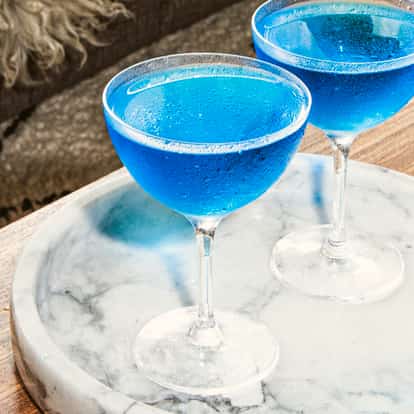 Coquetéis Blue Dolphin Martini em taças coupe geladas com azul vibrante, servidos em uma bandeja de mármore branco sobre uma mesa de madeira ao lado de um tapete.
