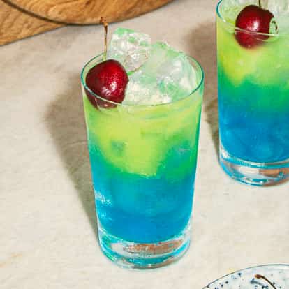 Blue Hawaiian Cocktails in hohen, gekühlten Gläsern mit blau-grünen Schichten, garniert mit Eis und Kirschen, serviert auf einer hellen Marmorarbeitsplatte.