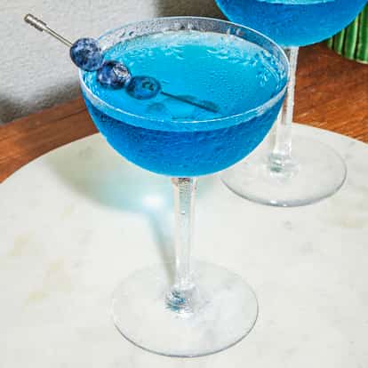 Gekühlte Blueberry Martinis in Coupette-Gläsern mit leuchtendem Blau, garniert mit Blaubeeren am Spieß, serviert auf einem runden Marmor-Tablett auf Holz.