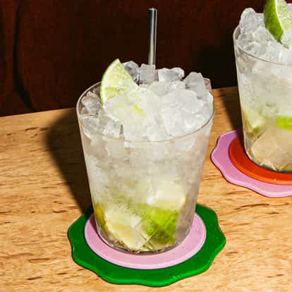Caipiroska cocktail in two ice-filled glasses with lime wedges and metal straws on colorful coasters on a light wooden table.
