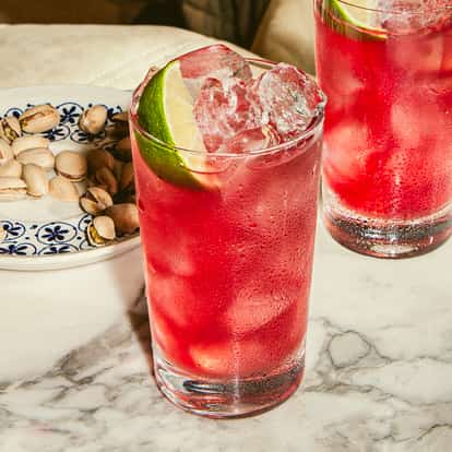 Dwie wysokie szklanki koktajlu Cape Cod z wódką, sokiem żurawinowym, lodem i kawałkami limonki na marmurowym stole, pistacje obok.