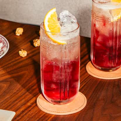 Dwie wysokie szklanki koktajlu Cosmow Mule z lodem, sokiem żurawinowym, ginger beer i plasterkami pomarańczy na drewnianym stole.