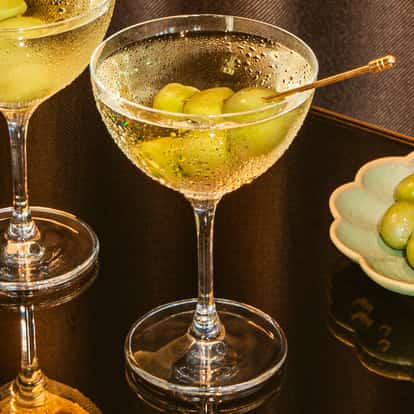 Two dirty vodka martinis with green olives in coupe glasses on a dark reflective table beside a plate of fresh green olives.