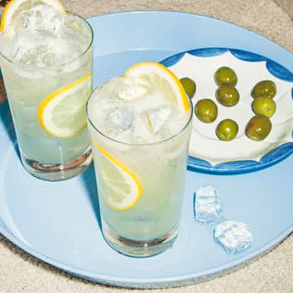 Elderflower Collins cocktails with lemon slices and ice on a blue tray beside a small dish of green olives on a carpeted surface.
