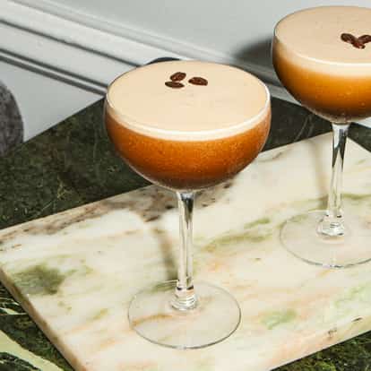 Two espresso martinis in elegant cocktail glasses on a marble table with roasted cashew nuts on a small plate.
