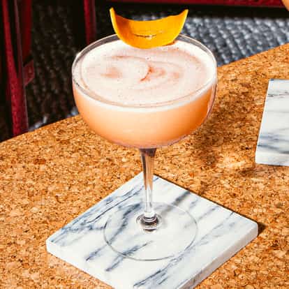 Cóctel Fancy Nancy en copa coupe con espuma cremosa y piel de naranja, sobre mesa de corcho con posavasos de mármol.