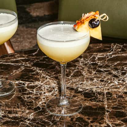 Flirtini-Cocktails in Coupégläsern, garniert mit Ananas und Kirsche, auf einem dunklen Marmortisch.