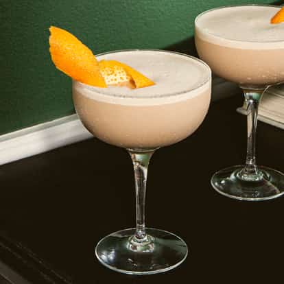 Galway Grey Cocktails in gekühlten Coupette-Gläsern mit cremigem Beigeton und Orangenzeste, serviert auf einer dunklen Oberfläche neben gestapelten Büchern und grüner Wand.