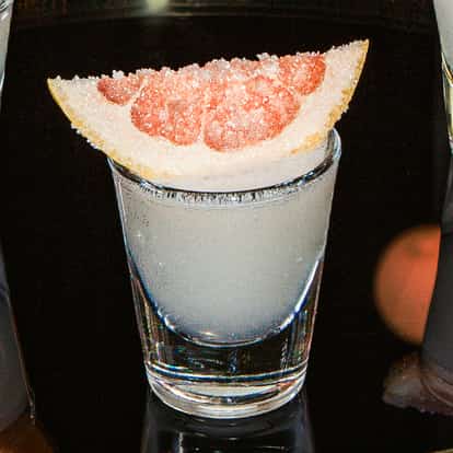 Grapefruit-Drop-Shot im klaren Shotglas mit einer Grapefruitspalte obenauf, auf einer glänzend schwarzen Oberfläche.