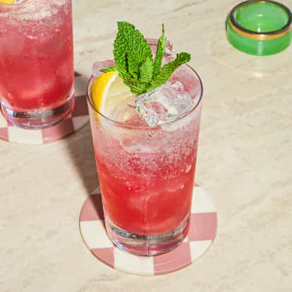 Zwei Grenadine-Fizz-Cocktails in hohen Gläsern mit Eis, Zitrone und Minze, serviert auf karierten Untersetzern auf einem hellen Steintisch.