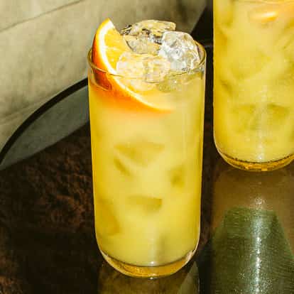 Drink Harvey Wallbanger w wysokiej szklance z lodem i plasterkiem pomarańczy, złoty koktajl na ciemnym szklanym stoliku.