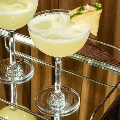 Hawaiian Cosmopolitan w kieliszkach coupe ze świeżymi plastrami ananasa na lustrzanej tacy na drewnianej powierzchni.