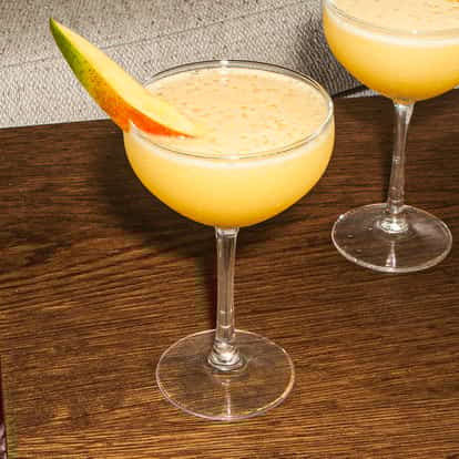 Mango No. 5 Cocktail in einer Coupette, cremig gelb mit einer Mangoscheibe, serviert auf einem Holztisch neben Büchern und einem Sofa.