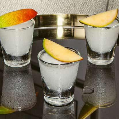 Mango Shooter in drei vereisten Shotgläsern, jeweils mit einer frischen Mangoscheibe garniert, präsentiert auf einem glänzenden schwarzen Tablett.