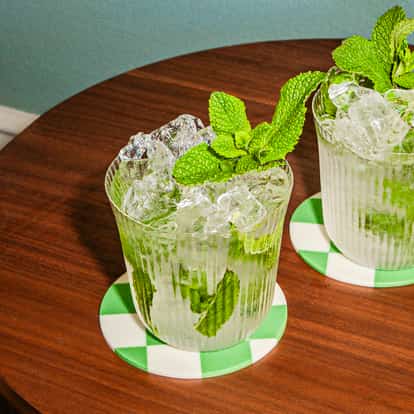 Dois coquetéis mint julep com gelo picado e hortelã, servidos em copos texturizados sobre porta-copos xadrez verdes.