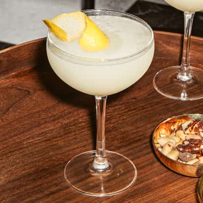 Nikita-Cocktails in eleganten Coupégläsern mit heller, schaumiger Oberfläche und Zitronenzeste, serviert auf einem Holztablett mit Nüssen und grünen Oliven.
