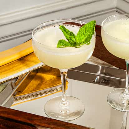 Dos copas coupe con cócteles espumosos y menta sobre una bandeja espejada junto a un bolso amarillo y una decoración verde.