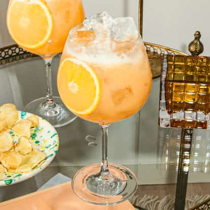 Koktajl Orang-a-tang w dużych kieliszkach na nóżce z lodem i plastrem pomarańczy, podany na stylowej tacy obok chipsów i ozdobnego szkła.