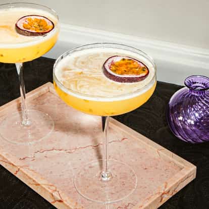 Due martini alla frutta della passione in coppette, guarniti con mezza frutto della passione su vassoio in marmo rosa.