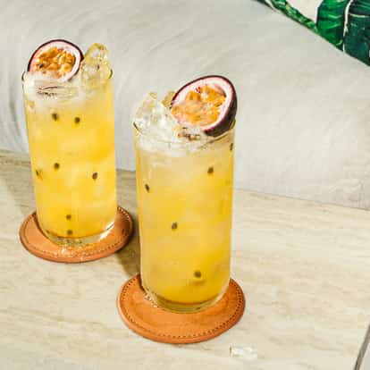 Wysokie szklanki z chłodzonym koktajlem z marakui na marmurowym stoliku, ozdobione połówkami owocu obok biżuterii i beżowej kanapy.