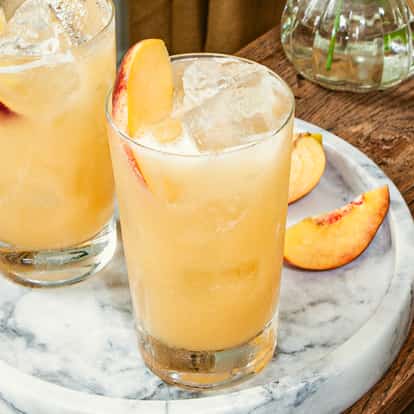 Peach on the Beach Cocktails in Highball-Gläsern mit frischen Pfirsichscheiben auf einem Marmortablett.